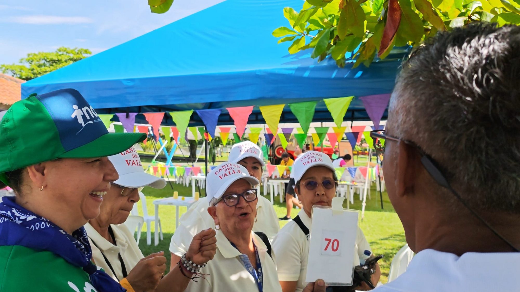 Adultas mayores con gorras del grupo Canitas del Valle conversando durante una actividad de los juegos, bajo una carpa azul con banderines de colores