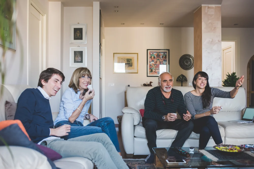 Familia de cuatro personas riendo en la sala de su casa mientras toman caf&eacute;